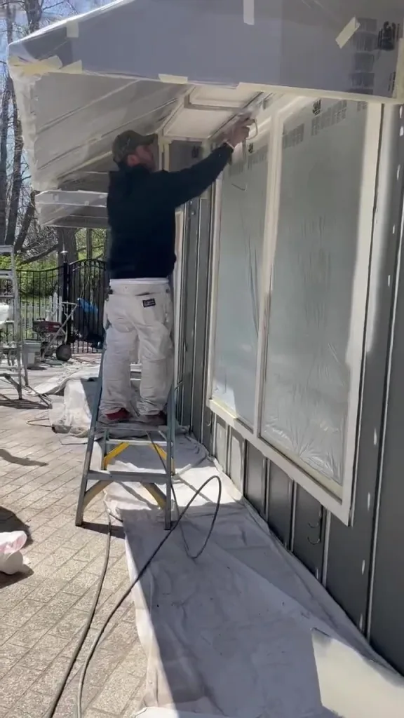 A painter in whites on a ladder, spraying the trim of a covered patio, drop cloths taped and spread below